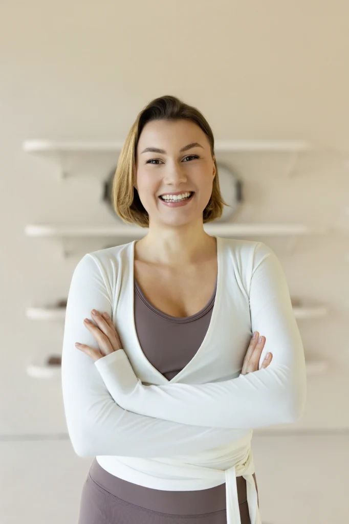Portret uśmiechniętej kobiety w sportowym stroju na tle jasnego studia pilates. Naturalne światło podkreśla przyjazny i profesjonalny wizerunek.
