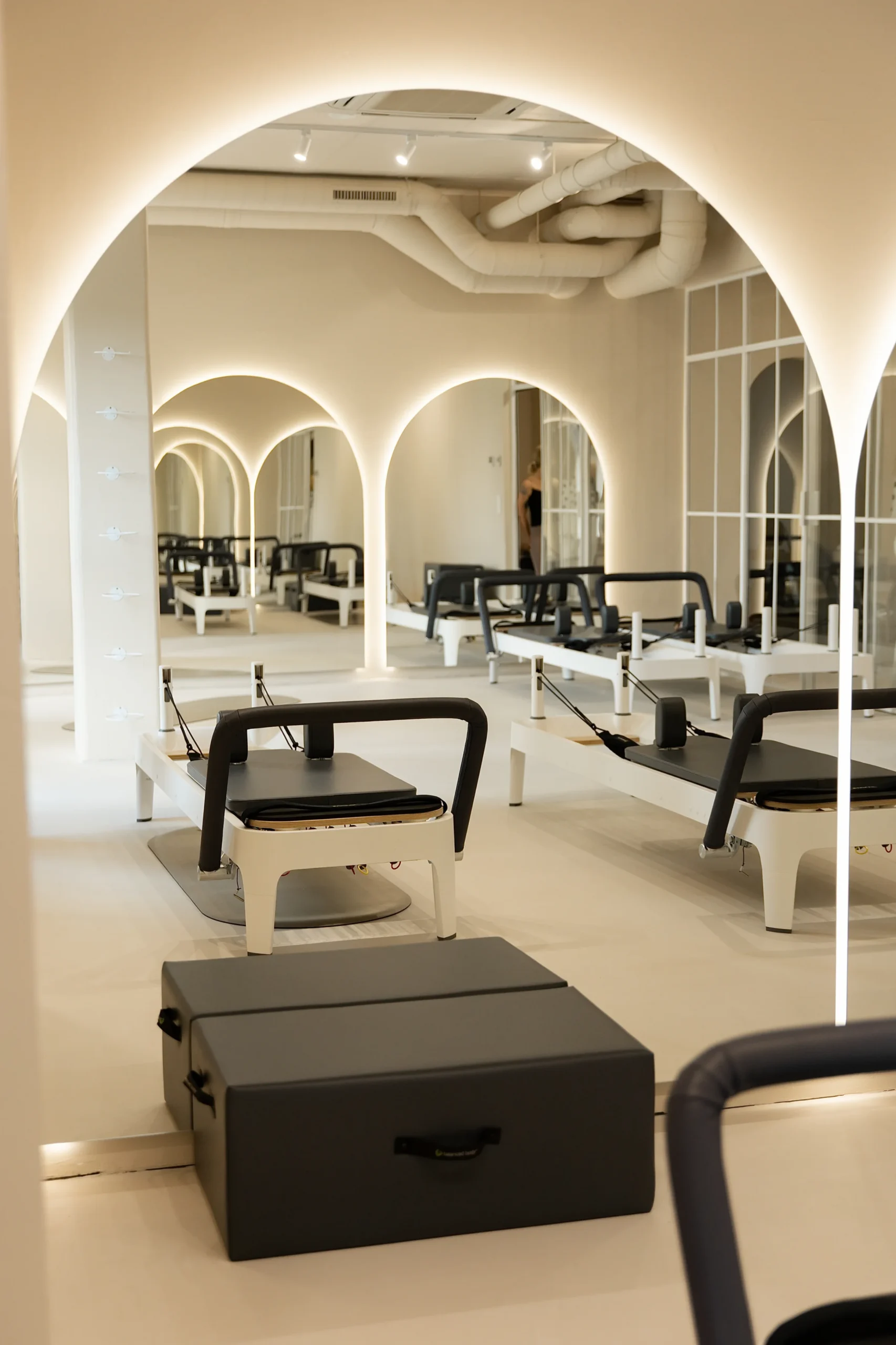 Reformer ustawiony w eleganckim studiu pilates z łukowymi przejściami i lustrami. Przestrzeń jest jasna, minimalistyczna i przygotowana do treningów indywidualnych.